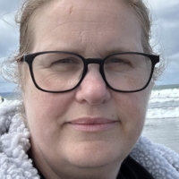 A woman with glasses smiles slightly at the seaside, with waves and cloudy sky in the background.