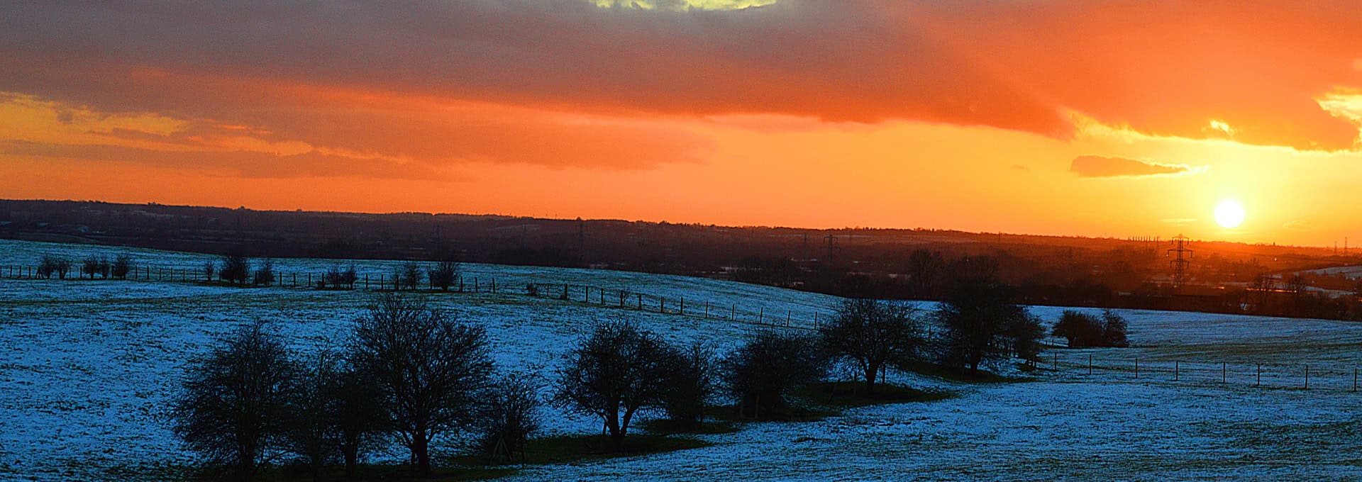 A snowy field with scattered trees at sunset, with orange and red sky on the horizon.