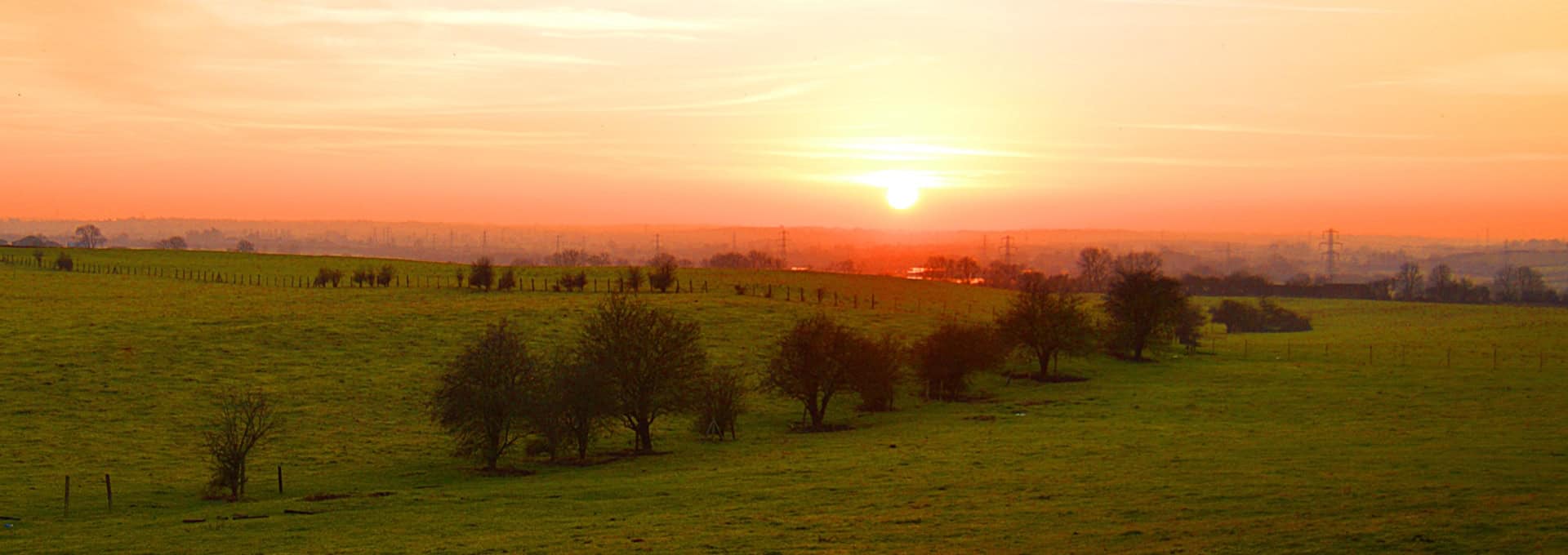 A bright orange sunset over a green field with scattered trees and a lightly clouded sky.